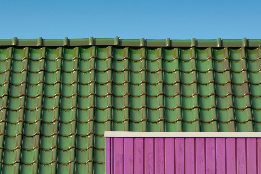 Roof Replacement A close-up of a green tiled rooftop against a blue sky with a contrasting pink wall.